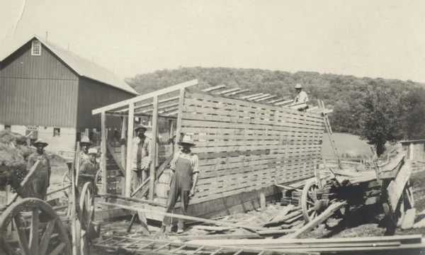 Photographic postcard of workmen posing with a partially built shed or corn crib. One man in sitting on the roof timbers. Two wagons with lumber are parked on two sides. A barn is in the background on the left, and wooded hills are in the far background.