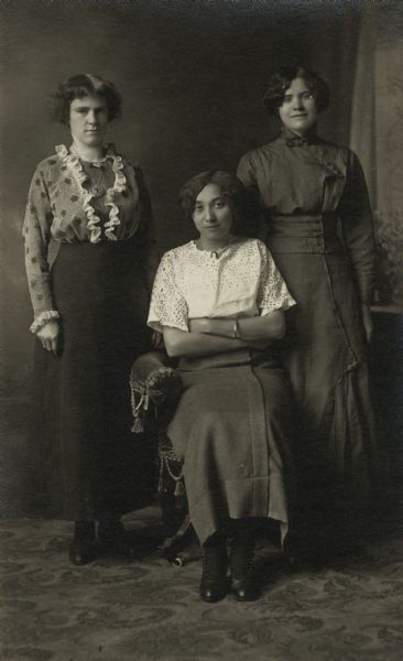 Photographic postcard of a studio portrait of three women, one seated in the center with her arms crossed and two standing behind her on both sides. The seated woman is Ann Drucella Witt, sister of Matthew Witt, the owner of the photography studio. In the background is a painted backdrop.