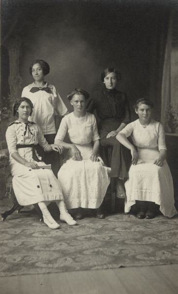 Portrait of five women, two standing and three seated in chairs. The woman standing on the left is Ann Drucella Witt, sister of Matthew Witt, owner of the photography studio. Two of the seated women are Agnes Marking and Elizabeth Juris.