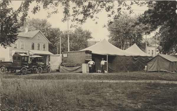 Photographic postcard of the Thompson Tent Show. Two tents, a tall wagon, an automobile and a truck are set up in a field or park with houses and trees in the background. Two men stand in the center with two small dogs at their feet. Painted on the side of the wagon, "Frank H. Thompson Big New Show." The tall wagon probably housed the movie projector.