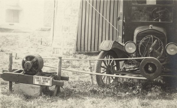 Photographic postcard of the generator used to power the "Frank H. Thompson Famous Moving Pictures." A belt from the generator stretches to the flywheel on the front of an automobile. Behind that is a billboard with many photos attached to it, probably advertising the movies being shown. In the background is a striped tent and a building.