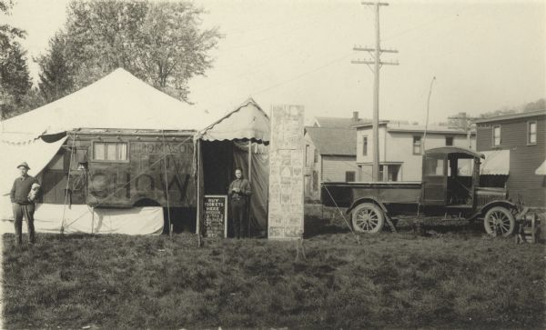Photographic postcard of the Thompson Tent Show. A tent, a tall wagon and a truck that runs the generator are set up in a field or park with houses and trees in the background. One man stands in the center next to a sign that reads, "Buy Tickets Here. Admission 22-TAX 3, Children 10 Cents Under [????] Years Old, Under Five FREE." On the other side is a billboard, with a poster advertising the film "The Birth of A Race," on top and 25 still photos below. Painted on the side of the wagon is "Thompson Tent Show." The tall wagon probably housed the movie projector. Another man stands on the left holding a small dog.
