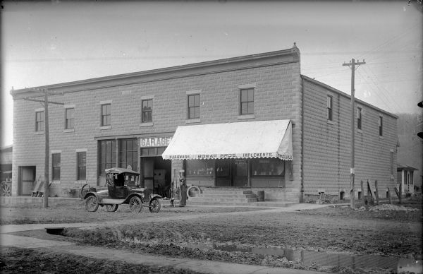 Photographic postcard of Bowar Bros. Hardware Store. The awning reads, "Hardware, Bowar Bros., Implements, Auto Supplies." Over the large door on the left is a sign, "Garage." An automobile and ladder can be seen inside. Steps lead up to the doors with display windows on both sides. Merchandise is on display in the windows and on the steps. An automobile is parked in the muddy street with two gas pumps at the curb.