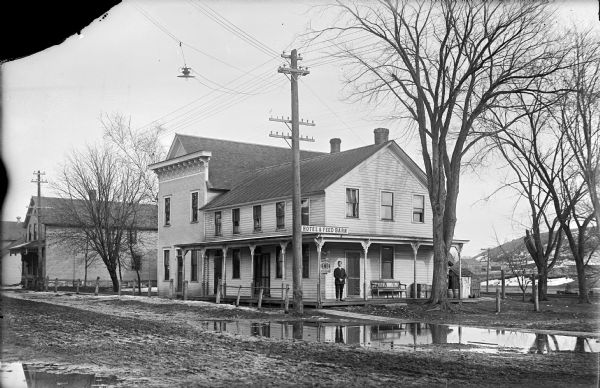 Photographic postcard of view across muddy road of a man wearing a suit and standing on the porch of the two-story Hotel and Feed Barn. Two signs next to him read, top, "BEER" and bottom, "Tom Moore 10¢ Cigars." The porch wraps around two sides of the building. On the right side are a bench, crates and barrels. Two more buildings are on the left. On the right are posts, railroad tracks and a snow-covered hill. Large trees surround the hotel.

