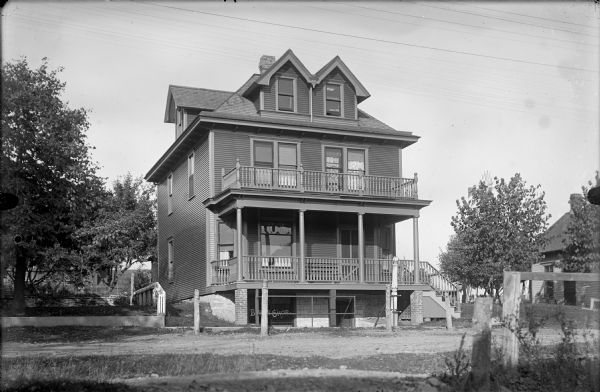 Front three-quarter view of large frame residence with porch and balcony. Steps with a barber pole at the top of the railing lead to a store window at foundation level, with the words: "Barber Shop" painted on the window.