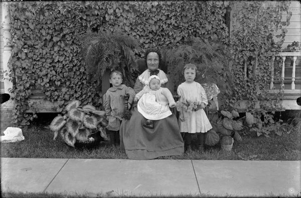 Group portrait of an elderly woman and three small children, including an infant. The woman is seated in a chair with the infant on her lap. A boy stands on the left, and a girl holding flowers stands on the right. Caladium in pots are arranged on both sides. In the background are ferns and a vine covered front porch. A sidewalk is in the foreground.