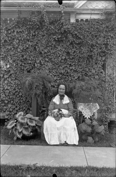 Portrait of an elderly woman. She is seated in a chair holding flowers on her lap. Caladium in pots are arranged on both sides. In the background are ferns and a vine covered front of a porch. A sidewalk is in the foreground.