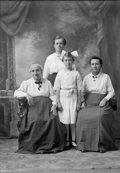 Studio portrait of three women and a girl in front of a painted backdrop. Two of the older women are seated, and the girl, wearing a large hair bow, is standing in between them. The other woman, who is wearing eyeglasses, is standing in the back.