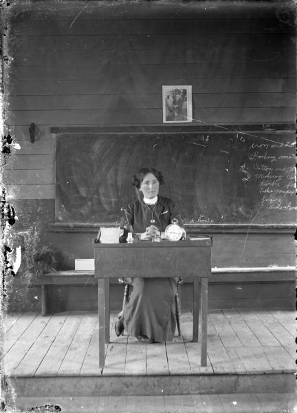 A schoolteacher sits behind her desk, on a raised platform, in a classroom. On her desk is a clock, inkwell, several books, hand bell and a hinged box. In the background is a blackboard. Above it hangs a portrait of Woodrow Wilson.
