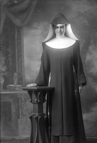 Three-quarter length studio portrait of a nun standing in front of a painted backdrop. She is resting one hand on a small pedestal table. She is wearing a habit, wimple and veil. A knotted cord shows at her waist on her right and a rosary and cross on her left.