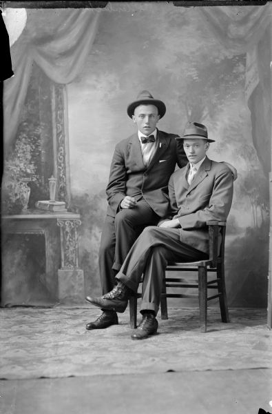 Formal full-length portrait of two men dressed in suits and hats. One man, wearing a necktie, is seated in a chair, and the other man, wearing a bow tie, is perched on the arm of the chair with his arm draped over the back.