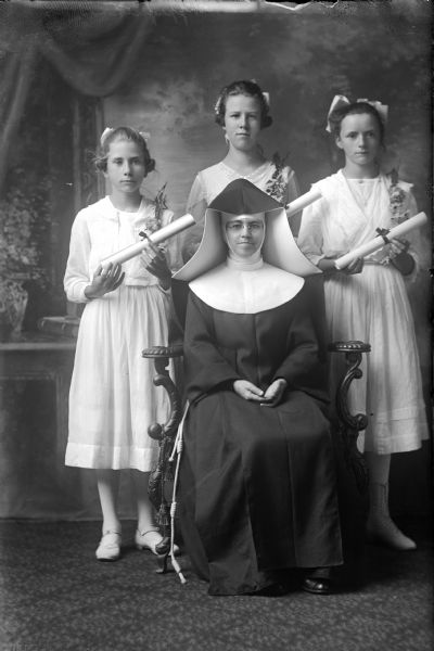 Full-length studio portrait in front of a painted backdrop of a nun seated in a chair and three girls posed behind her. The nun is wearing a habit, wimple and veil and the girls are wearing white dresses, stockings, shoes and hair bows. The girls are each holding a scroll tied with a ribbon and have a corsage pinned to the shoulder of their dresses.