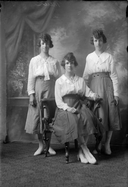 Full-length studio portrait of three young women posed in front of a painted backdrop. The middle woman is seated and the others stand on each side behind her. All three are dressed alike in dark matching skirts, light blouses, white lace-up shoes and jewelry. They also have matching hairstyles and may be sisters or cousins.
