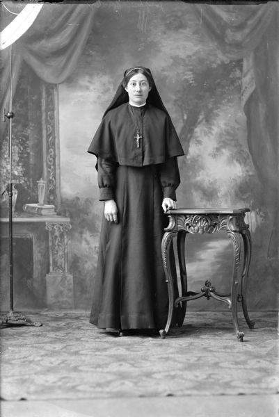 Full-length studio portrait in front of a painted backdrop of a nun, Josefine Zander. She is wearing a habit and veil with a crucifix around her neck. Her left hand rests on an ornate table. On her right, the studio light stand can be seen.