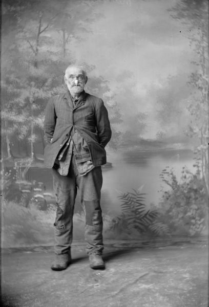 Photographic postcard of a full-length studio portrait in front of a painted backdrop of a man standing with his hands behind his back. He is wearing work clothes and is smoking a pipe. The pipe is partially obscuring his face.