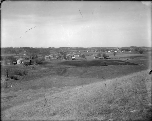 View from grassy hill of uptown Cross Plains. The Catholic Church (St. Francis Xavier) is on the right, and farmhouses and other buildings are among fields. In the far background are more hills.