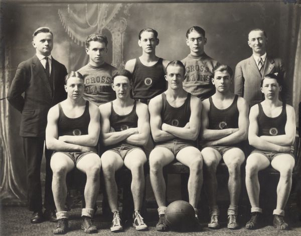 Studio portrait of the Cross Plains American Legion basketball team, coaches and assistants in front of a painted backdrop. The players wear uniforms, the coaches wear suits and the assistants wear sweatshirts with "Cross Plains" embroidered on them. The front row of players are seated and have their arms crossed. A basketball rests on the floor in the foreground. Their names are (back row, l to r) Tom Daniels, Eddie Schmitz, Pinney Faust, Roman Esser and August Stumpf, (front row, l to r), Bubby Vasen, Jack Hammerli, Harold Saether, Hank Bollig and Hardy Endres.