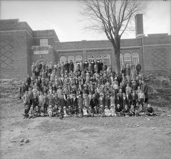 Large group of students and teachers posed on a slope in front of St. Francis Xavier Catholic School for a group portrait. Many of the children and adults are wearing coats and hats, and a tree on the slope does not have leaves.