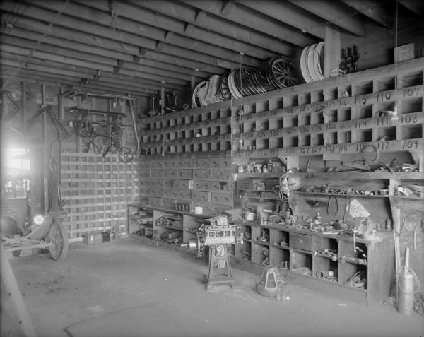 Interior view of the Bowar Bros. Garage. On two walls are banks of drawers and cubby holes for tools and parts. On the right are more shelves for tools. Among the tools are metal shears, drills, wrenches, hacksaws, a vise and oil cans. Wheels are stored near the ceiling on the right. More parts hang on the wall on the left. The front of an automobile is parked at the garage door. Electric lights hang to illuminate the work area.