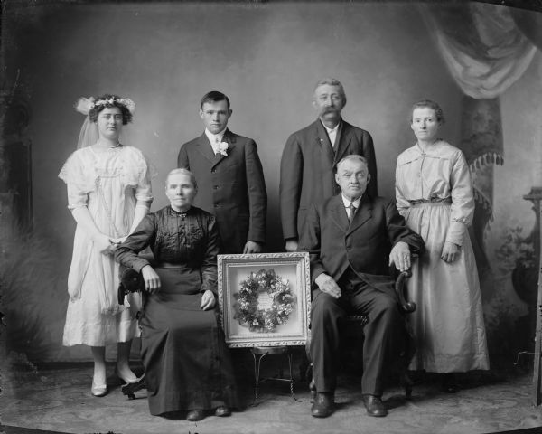 Studio family wedding portrait in front of a painted background with a framed wreath in the center. The wreath is celebrating the wedding of the bride and groom in the back on the left, the 25th anniversary of the couple (parents of the bride) on the right and the 50th anniversary of the seated couple (parents of the groom) in the front. The text inside the wreath reads, "Paul und Mina Ketelboeter, 1867--12. Dez.--1917. [????] und Hulda Marten, 1892--12. Dez--1917. Percy und Mina Ketelboeter, 1917--12. Dez.--1917."