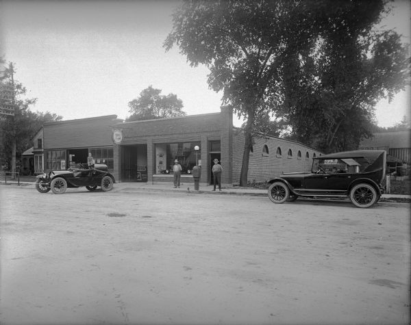 View across road of D.A. Meister's Garage, with a fire truck parked in front and an automobile parked at the curb on the right. One boy is sitting on the fire truck and another child peers out of the automobile. Two men pose on either side of a street light, in front of the display window, with what may be a call box on it. One or more automobiles are on display inside. Hanging over the garage door is a round "Authorized Buick Service" sign with two lights aimed at each side. Behind the garage door is part of a sign for "Meister Buick Motor Cars." "D.A. Meister Garage" is painted on the display window. Looking through the parked car, a sign for "Camel Cigarettes" and "Holsum Bread" can be seen on a small shed, and to the right may be a barn. A dwelling is on the left.