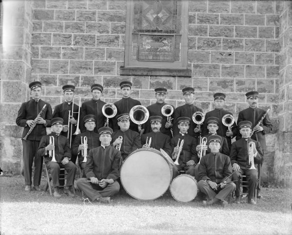 Outdoor group portrait of the band of St. Martin's Catholic Church. The members pose with their instruments, which include clarinets, french horns, flugelhorns, baritone, trombone and drums. Brickwork and a stained glass window are behind them.