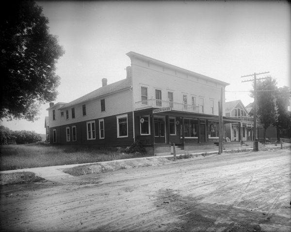 Undertaker's funeral parlor and other buildings along a dirt street with a sidewalk. An oblong sign attached to the balcony hangs over the sidewalk and reads, "Undertaker." Two round signs advertising a telephone hang under the portico. Displayed in the windows on the right is furniture. A dwelling is on the far right.