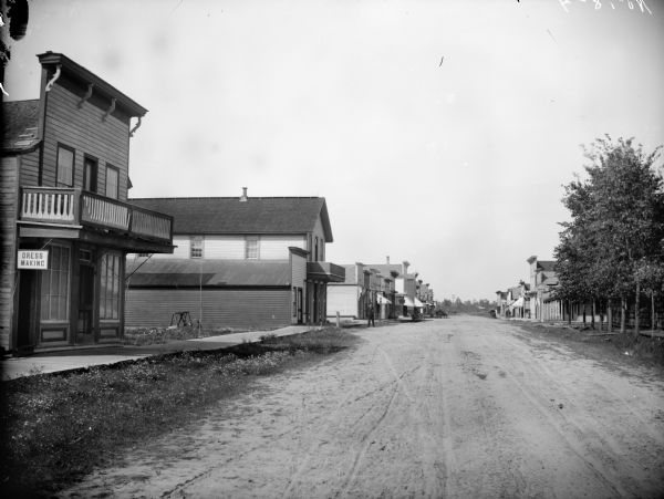 View down dirt road of buildings and storefronts along a sidewalk, possibly Main Street, looking north toward the Chicago & Northwestern railroad tracks. A man is standing on the street, and there are signs for the American House, Dress Making, and Tin Shop.