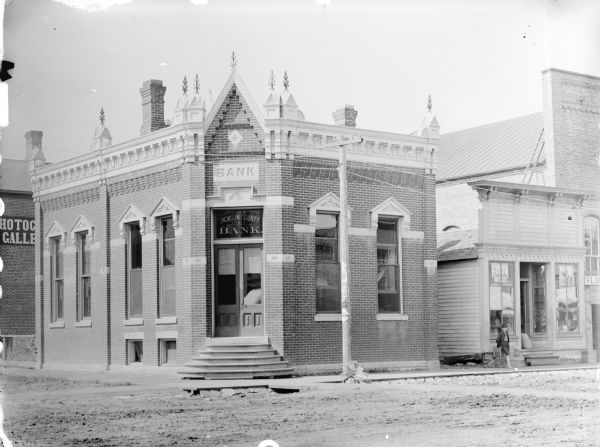 Jackson County Bank and the Norse Handel Grocery Store on the corner of First and Main Streets. A young boy is standing on the sidewalk in front of the grocery store. The Van Schaick Photography Gallery is partially visible on the left.