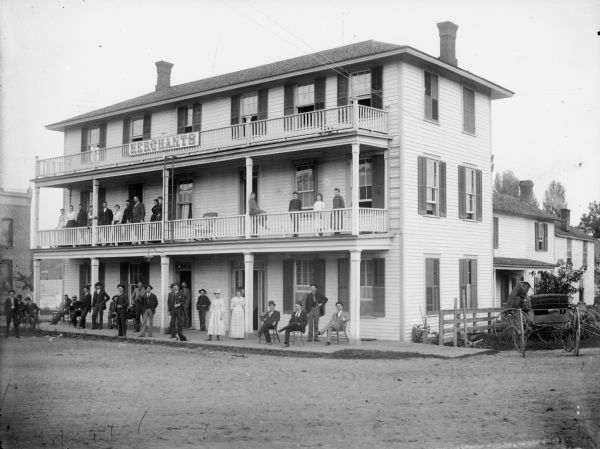 View from street of the Merchants Hotel, located at the intersection of First and Fillmore Streets. A large group of men and women, and a puppy, are posed standing and sitting on the ground and second floor porches.