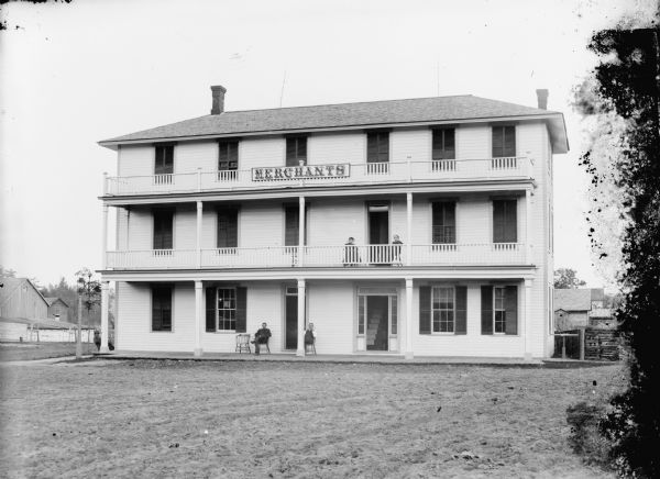 View of front of Merchants Hotel, located at the intersection of First and Fillmore Streets. Two men are posed sitting on the first floor porch, and two women are posed sitting on the second floor porch.