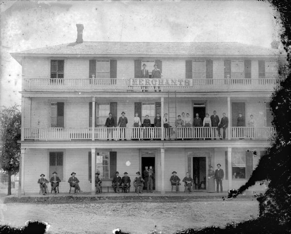 Exterior view of the facade of Merchants Hotel, located at the intersection of First and Fillmore Streets. Men and women are posed sitting and standing on the porches of all three floors.
