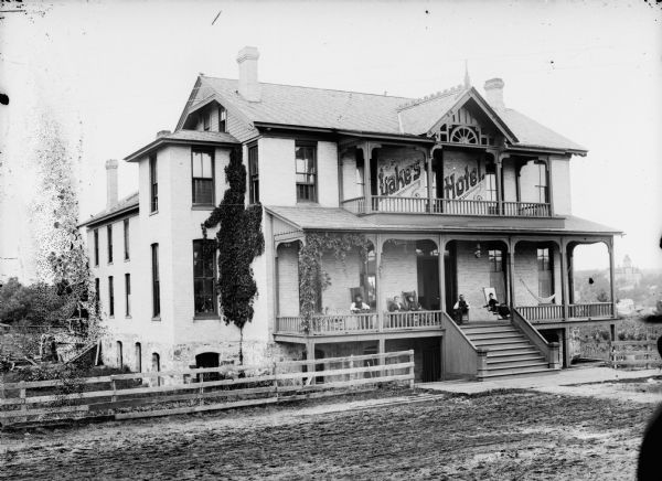 Exterior of Lake's Hotel with people posing sitting on the lower porch. Possibly located across from the railroad depot across the Black River, probably in the center of the block where the Emma Gerhardt House later stood. The hotel burned down and the cream-colored brick was used to construct the Waughtal Mill.