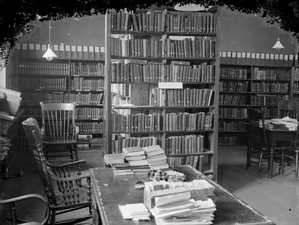 Interior of a library with tables and shelves full of books, probably the Black River Falls Library located in a building next door to the house of Frances Perry, later burned in 1917 or 1918. After the flood of 1911, dances were held in the library and Mrs. Merlin Hull played an organ.