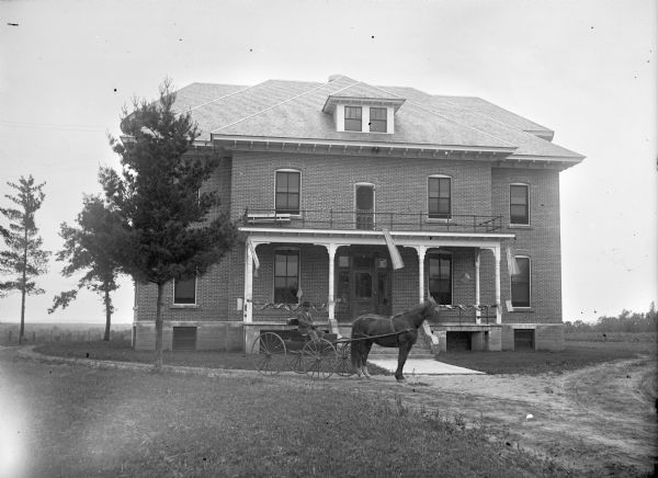 Man posed sitting in a buggy pulled by a single horse in front of a two-story brick building, probably the Jackson County Poor Home.
