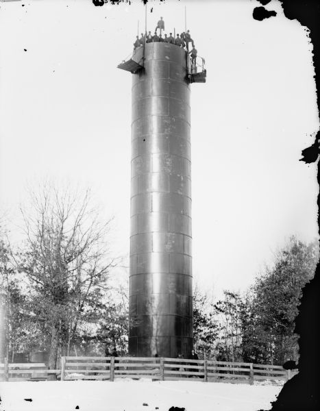 Men posed standing on top of the City Water Tower, probably seventy feet high. A smaller group of men stand at the base behind a fence, and there is snow on the ground.