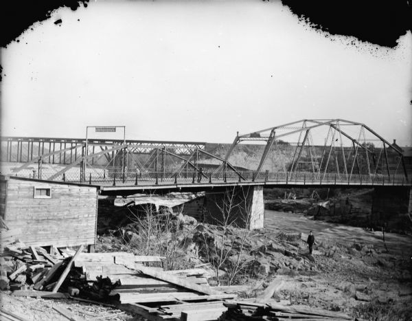 Men posed standing on the rocky river bank and bridge, probably the Harrison Street Bridge. A sign on the bridge says: "? Fine for driving or riding on this bridge faster than a walk." There is a wooden structure on the left with lumber stacked on the ground nearby.