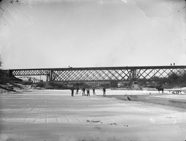 Six men harvesting ice, probably from above the river dam on the Black River, in front of the railroad bridge.	There is a horse standing on the ice on the right, and people are standing on the railroad bridge.