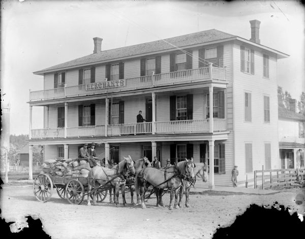 View from street of two men posed sitting in a wagon full of grain bags and pulled by a team of four horses in front of the Merchants Hotel. L.S.Y. June, the hotel proprietor, is probably the person standing without a hat behind the lead horses.