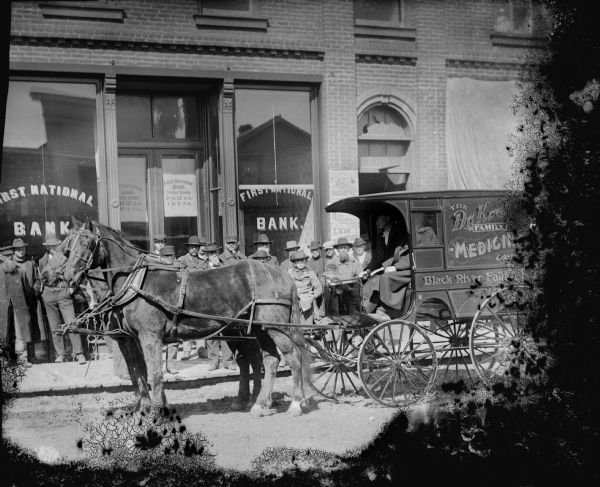 View from street of Dr. Krohn Family Medicine Wagon pulled by a team of two horses in front of a crowd of people posed standing in front of the First National Bank on Main Street.	
