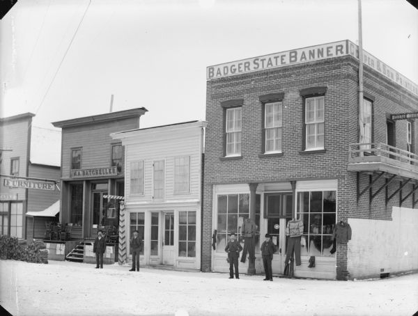 View across street of four men posing in front of storefronts on Main Street, including M.A. Batcheller, a barber shop, and clothiers under the offices of the Badger State Banner. There is snow on the ground. Fuelwood is stacked on the sidewalk on the left.