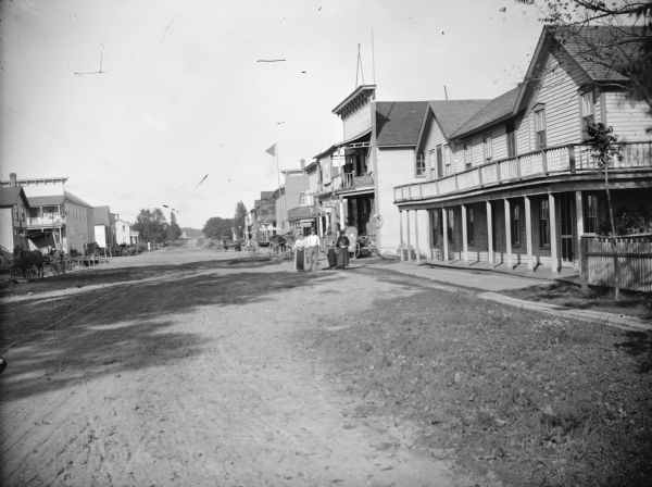View down street lined with storefronts, probably Alma Center. People are standing and posing near storefronts. Low building in the right foreground in probably the Clark Hotel, and the M.J. Chapman Store is across the street. The right side of the street was later destroyed by a fire.	