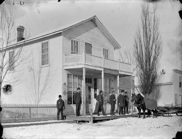 Men posed standing in front of the S.H. Van Gorden & Son General Merchandise Store on a snow-covered street. S.H. Van Gorden is probably standing on porch at left. Two horses on the right a pulling a sled. The store burned down 1903.	