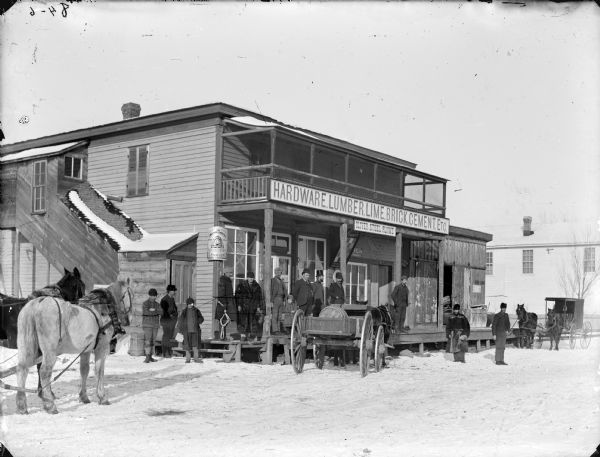 Group of men and boys posed outside the Miller Store. There is snow on the ground. The photographer's wagon pulled by two horses is at the left.	