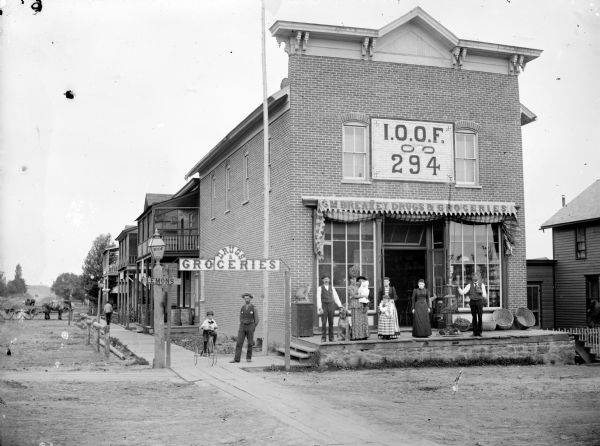 Group of men, women, children and a dog posing standing in front of G.M. Breakey Drugs and Groceries. One man, perhaps the grocer, is standing in front of the open doorway holding a bottle in one hand, and a glass of liquid in the other. A small child is riding a tricycle. Baskets of fruit and or vegetables are on display outside the storefront. The second floor of the building is the Masonic temple for International Order of Odd Fellows, Lodge 294.	