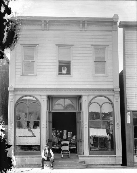 View from street of the Nick Nelson Furniture Store, with a man posed sitting on the steps. A rocking chair sits in the middle of the stoop in the open doorway.