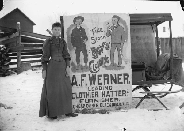 Woman posing outdoors standing next to an advertisement for the A.F. Werner Store, which is attached to a sleigh on a snow-covered street. The large poster depicts two boys, one of whom is holding ice skate blades, flanking the text: "Fine Stock of Boys Clothing."