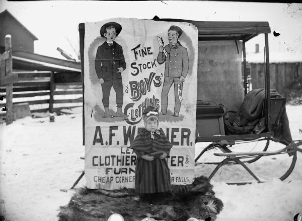 Small girl posed outdoors on a fur rug in front of an advertisement for the A.F. Werner Store. The poster is attached to a sleigh on a snow-covered street, and depicts two boys, one of whom is holding ice skate blades, flanking the text: "Fine Stock of Boys Clothing."