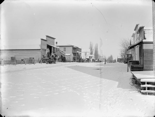 View of a street in a town, probably Hixton, with footprints crisscrossing a snow-covered street. A group of men are standing on the porch of the hardware store, and horses and carriages are parked along the street.
