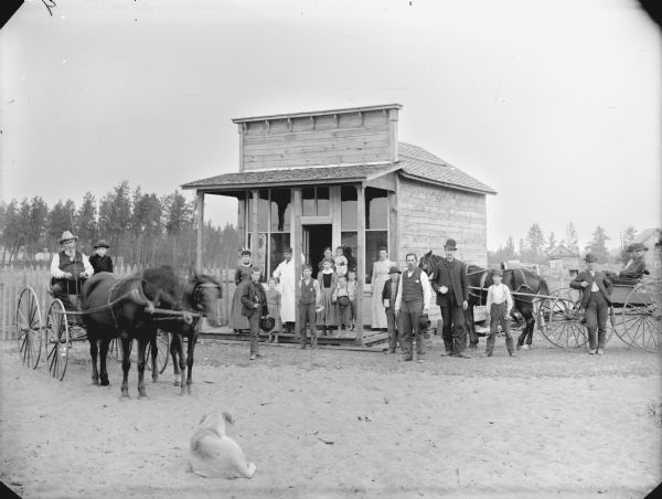 Group of men, women, and children posing standing in front of a small country store. A man and boy are sitting in a buggy pulled by a single horse. In the foreground a dog is sitting in the road.